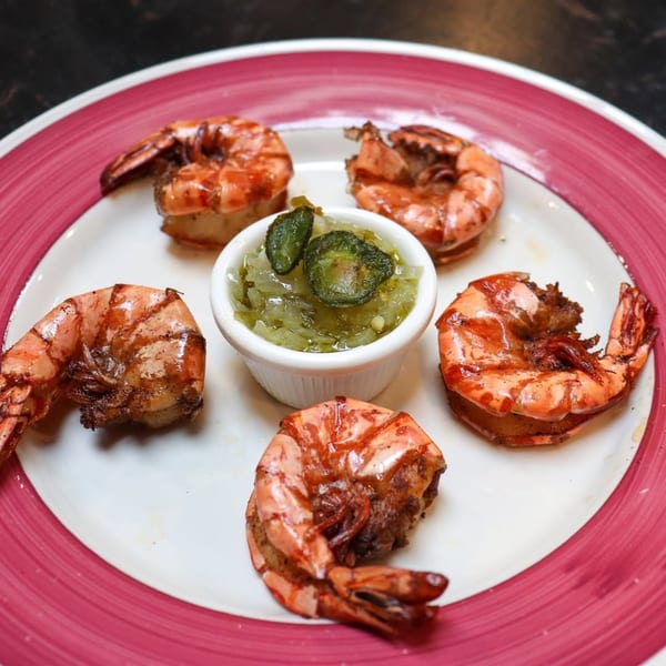 Five grilled shrimp arranged around a small white bowl of green relish on a white plate with a red rim.