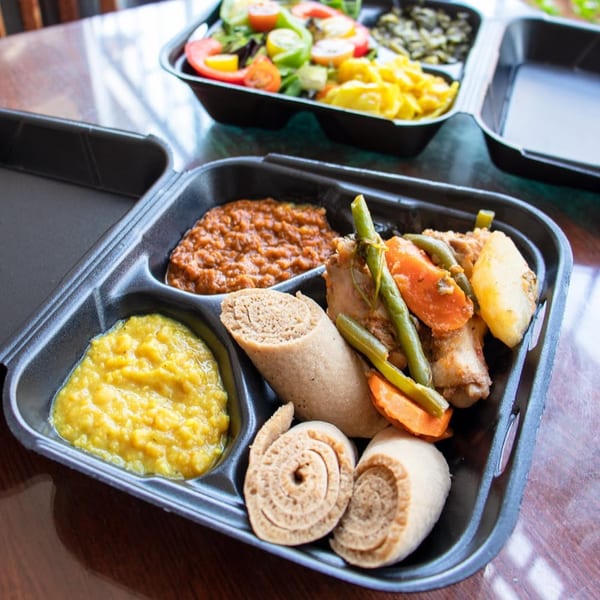 Ethiopian takeout meal with injera, stews, vegetables, and a side salad in black containers.